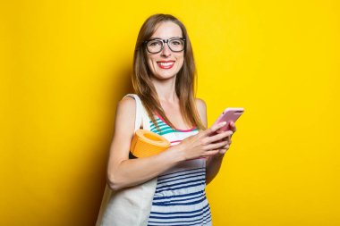 Sarı arka planda yoga minderi olan, omuz çantanda telefonla gülümseyen arkadaş canlısı genç bir kadın.