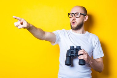 Şok olmuş sakallı genç adam elinde dürbünle sarı arka planı işaret ediyor..