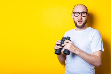 Şok olmuş sakallı genç adam sarı arka planda dürbün tutarken kameraya bakıyor..