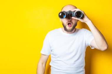 Sakallı genç adam sarı arka plandaki dürbünle şaşırmış görünüyor..