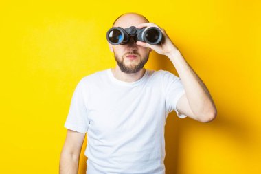 Sarı arka planda dürbünle bakan sakallı genç bir adam..