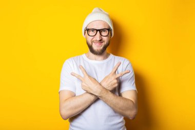 Şapkalı ve gözlüklü gülümseyen genç adam elleri ve kolları çapraz ve iki parmağı havada jestler gösteriyor.