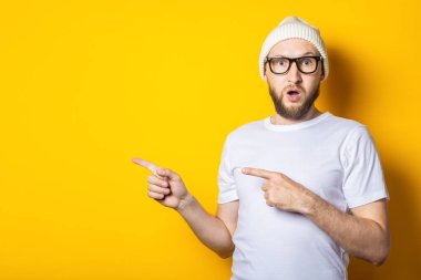 Şapkalı, gözlüklü, sakallı, şaşırmış genç adam sarı arka planda parmaklarını işaret ediyor.