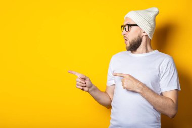 Şapkalı, gözlüklü, sakallı, şaşırmış genç adam sarı arka planda parmaklarını işaret ediyor.