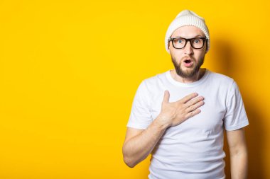 Şaşırmış sakallı genç adam sarı arka planda elini göğsüne koydu.