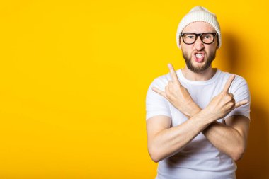 Çapraz kollu sakallı genç adam sarı arka planda rockçı bir keçi hareketi gösteriyor..