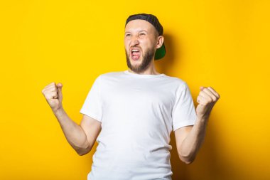 Mutlu sakallı, beysbol şapkalı, sarı arka planda yumrukları olan genç adam.