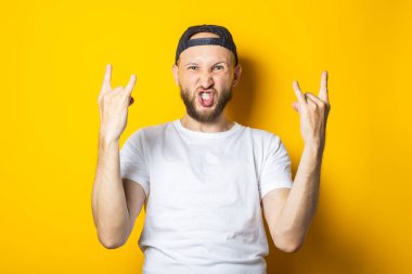 Bağıran sakallı genç adam sarı arka planda rockçı keçi pozu veriyor.