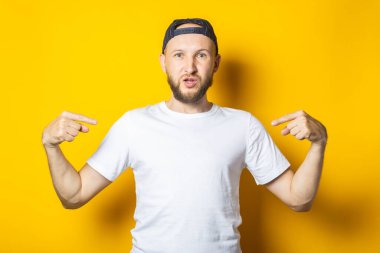 Beyaz tişörtlü, yazılı olmayan sakallı genç adam, sarı arka plandaki parmaklarıyla kendini işaret ediyor.