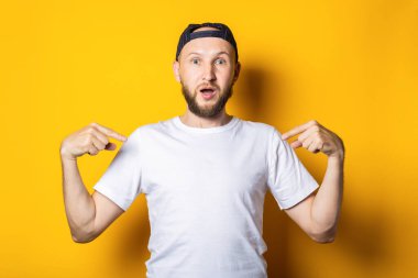 Beyaz tişörtlü şaşkın genç adam, kendini parmaklarıyla gösteriyor, mesaj için yer var, sarı arka planda.
