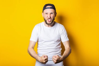 Sakallı, yumruklarını sıkmış, sarı arka planda erkeksi bir güç gösteren genç bir adam.