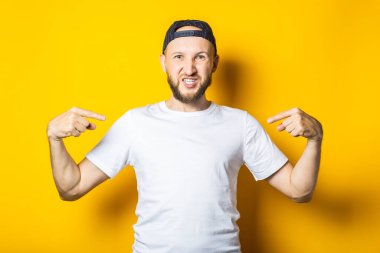 Beyaz tişörtlü, yazılı olmayan gülümseyen genç adam sarı arka planda parmakları olduğunu gösteriyor.