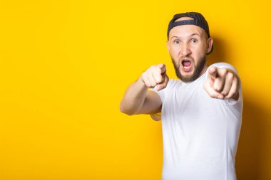 Beyaz tişörtlü genç adam sarı arka planda parmaklarıyla onu işaret ediyor.