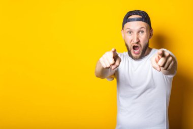 Beyaz tişörtlü sakallı genç adam bağırıyor ve sarı arka planda kamerayı işaret ediyor.