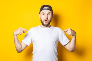 Beyaz temiz tişörtlü, sakallı, şaşırmış genç adam sarı arka planda kendini işaret ediyor.
