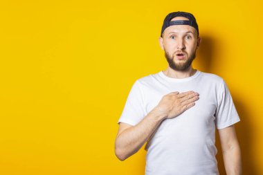 Sakallı genç adam sarı arka planda elini göğsünde tutuyor.