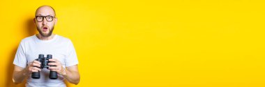 Sarı arka planda dürbün tutan gözlüklü sakallı genç adam şaşırdı. Pankart