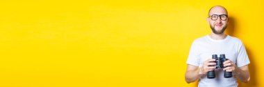 Sarı arka planda dürbünü tutan sakallı, gülümseyen genç bir adam. Pankart