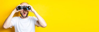 Şaşırmış sakallı genç adam sarı arka plandaki dürbünle bakıyor. Pankart