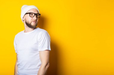 Beyaz tişörtlü, sarı arka planda gözlüklü, şapkalı sakallı genç adam.