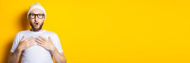 Şaşırmış sakallı genç adam sarı arka planda ellerini göğsünde tutuyor. Pankart