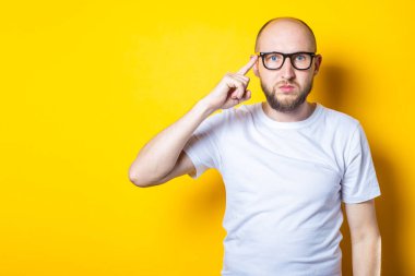 Ciddi sakallı, gözlüklü genç adam sarı arka planda şakağına parmağını uzatıyor.