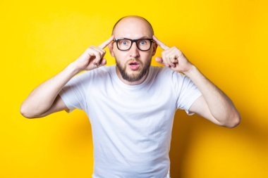 Ciddi sakallı, kel, gözlüklü genç adam sarı arka planda parmaklarını şakağına tutuyor.