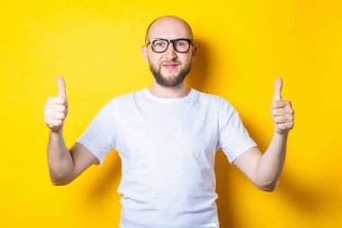 Gülümseyen sakallı genç adam elleriyle, başparmağıyla, sarı arka planda el hareketi yapıyor.