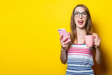 Neşeli bir şekilde şaşırmış genç bir kadın, elinde bir fincan kahveyle sarı arka planda..