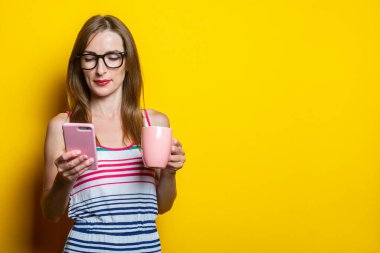 Odaklanmış telefonlu genç bir kadın, elinde kahve fincanı, sarı arka planda.