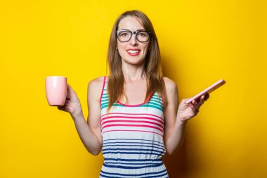 Elinde bir telefon ve sarı arka planda kahve olan güzel, gülümseyen bir kadın..