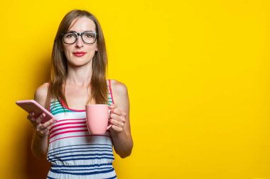 Güzel, dost canlısı bir genç kız elinde kahve ve sarı arka planda bir telefon tutuyor..