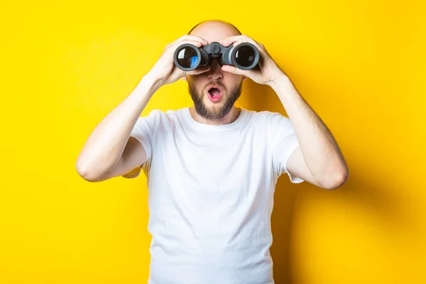 Surprised shocked bearded young man looks through binoculars on a yellow background.