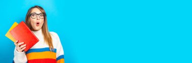 Şaşırmış yüzlü genç bir kadın mavi arka planda kitapları elinde tutuyor. Eğitim kavramı, üniversite, seans, sınav, kariyer seçimi. Pankart.