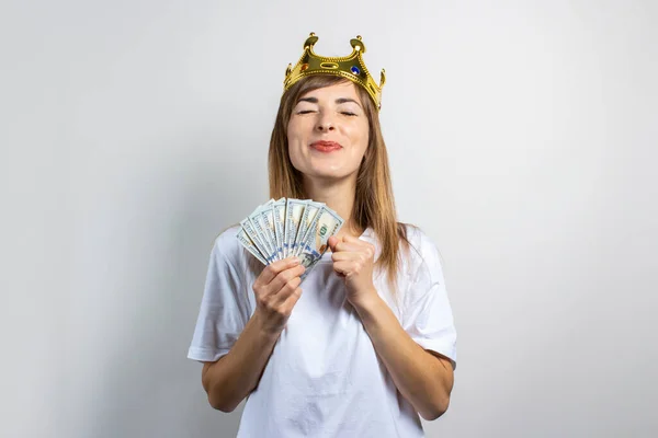 Young woman with a crown on her head holds a stack of money and celebrates very joyfully on a light background. Emotion laughter, surprise, kiss, shock. Queen, luck, wealth, gain, victory, bet. Banner.