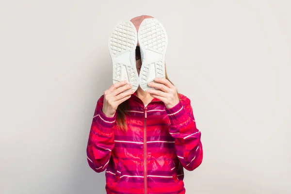 Young woman with glasses, hat and pink sports jacket with serious face covers her face with sneakers on a white background with copy space. Concept modern trendy style. Face expression. Banner.