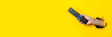 Man's hand holding a TV remote control on a bright yellow background. Banner clipart