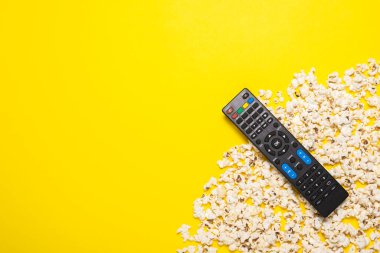 TV uzaktan kumandası, TV ayarlayıcısı ya da ses sistemi ve sarı arka planda patlamış mısır. Kavram serisi, film, spor. Pankart. Düz yatış, üst görünüm.