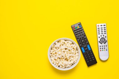 Televizyondan iki uzaktan kumanda, televizyon ayarlayıcısı ve sarı arka planda bir kase patlamış mısır. Kavram serisi, film, spor. Pankart. Düz yatış, üst görünüm.