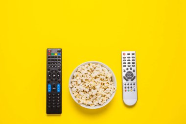 Televizyondan iki uzaktan kumanda, televizyon ayarlayıcısı ve sarı arka planda bir kase patlamış mısır. Kavram serisi, film, spor. Pankart. Düz yatış, üst görünüm.