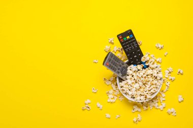 Televizyondan iki uzaktan kumanda, televizyon akortçusu sarı arka planda patlamış mısırla bir kasede yatıyor. Kavram serisi, film, spor. Pankart. Düz yatış, üst görünüm.