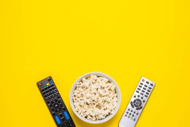 Televizyondan iki uzaktan kumanda, televizyon ayarlayıcısı ve sarı arka planda bir kase patlamış mısır. Kavram serisi, film, spor. Pankart. Düz yatış, üst görünüm.