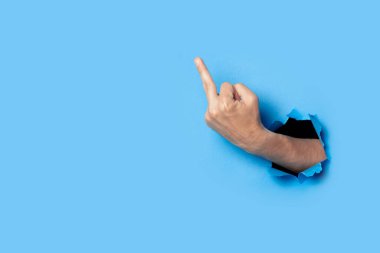 Hand shows the gesture of the middle finger on a blue background. Offensive gesture, fuck off, leave me alone.