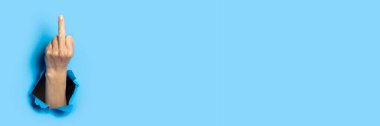 Hand shows the gesture of the middle finger on a blue background. Offensive gesture, fuck off, leave me alone.