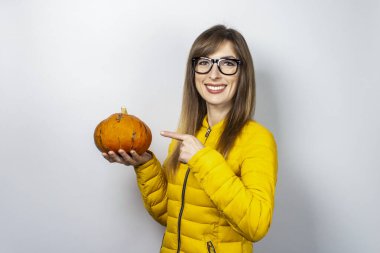 Sarı ceketli genç kız bir balkabağı tutuyor ve arka planda onu işaret ediyor. Cadılar Bayramı konsepti, sonbahar, kutlama.