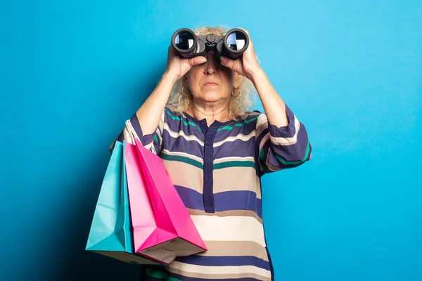 Stara Kobieta Trzyma Torby Zakupy Patrzy Przez Lornetkę Niebieskim Tle — Zdjęcie stockowe