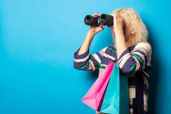 Old woman holding shopping bags and looking through binoculars on blue background.