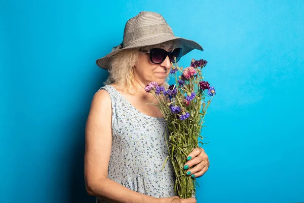 Old woman in a wide-brimmed hat and dress sniffing a bouquet of flowers on a blue background.