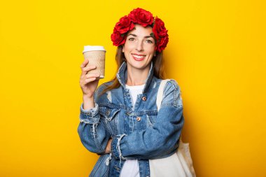 Kot ceketli bir gülümseme ve kafasında kırmızı bir çiçek çelengi olan genç bir kadın sarı arka planda kahve olan bir kağıt bardak tutuyor..