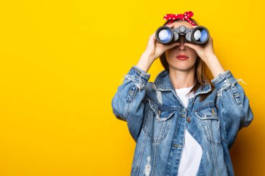 Kot ceketli ve kafasında bir sal olan genç kadın sarı arka plandaki dürbünle bakıyor. Pankart.